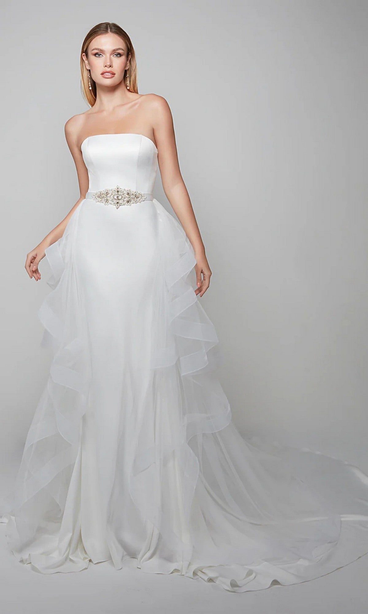A woman stands against a plain background wearing the Alyce 7025, a long white strapless satin dress with a fitted bodice, jeweled belt, ruffle-layered tulle overskirt, and a long train.